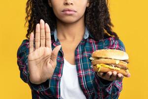 Say No To Fast Food. Closeup Portrait Of Serious African American Woman Holding Burger And Showing Stop Gesture With Palm. Black Lady Rejecting Unhealthy Meal, Isolated Over Orange Background, Banner photo