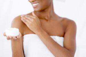 Advertising of cosmetic product for body skin moisturizing and anti aging. Young happy cute african american female in white towel shows cream in bedroom interior on bed, cropped, empty space photo
