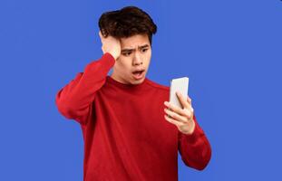Surprised Asian Young Man With Cellphone Reading Bad News Message Touching Head In Shock Standing On Blue Studio Background, Wearing Red Sweater. Confused Mobile Phone User, Smartphone Problem Concept photo