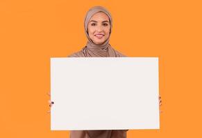 Muslim Lady Holding Blank Poster Board For Text Advertisement Posing Over Yellow Studio Background, Wearing Hijab Headscarf. Islamic Woman Advertising Something With Empty Paper Sheet Banner. Mockup photo