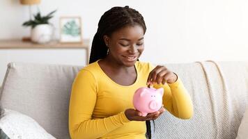 Young black woman putting coin in piggy bank while sitting on couch at home, saving money for future needs and utility payments, making wise financial planning and investments, copy space photo