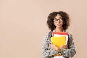 Human natural emotions of sadness and fatigue. Tired african american student in glasses with backpack hugs notepads and looks up at empty space, isolated on yellow background, studio shot, cut out photo