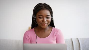 Smiling african american woman in headset watching webinar on laptop at home, enjoying online education and distance learning, sitting on couch and looking at computer screen, closeup with free space photo