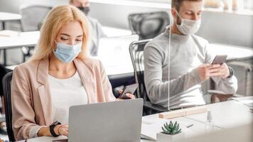 Modern gadgets for working with clients in office. Millenial man in mask typing on smartphone, lady looks at laptop at workplace with protective glass during covid-19 epidemic, empty space photo