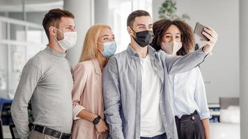 Corporate selfies, team building and new normal during COVID-19 outbreak. Multiracial millennial workers in protective masks makes photo with smartphone for work blog in interior of modern office