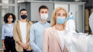Temperature control test at work and social distancing. Man in protective suit checks multinational workers with non-contact thermometer in morning at entering in office, empty space photo