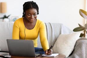 Online Education Concept. Smiling Black Woman In Headset Studying With Laptop At Home And Taking Notes To Notepad, Enjoying Distance Learning, Watching Webinar In Internet And Noting Highlights photo