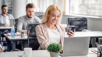 Manager works with client online and social distance. Smiling woman worker holding smartphone and typing on laptop at workplace in modern office interior returning after quarantine, copy space photo