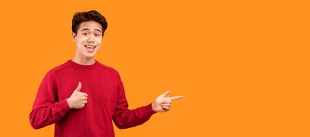 Look There. Smiling asian guy pointing his fingers away at copy space for your text or logo, isolated over orange studio background wall, banner, panorama. Smiling man showing thumbs up photo