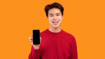 I Recommend This App. Smiling asian guy holding smartphone with black empty screen in hand and showing it to camera. Gadget with blank space for mock up, isolated over orange studio background photo