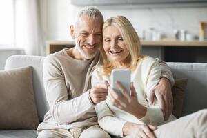 A happy senior couple sits on a couch, smiling as they share a moment while looking at a smartphone. Their expressions reflect joy and connection, showcasing their bond in a cozy living room. photo