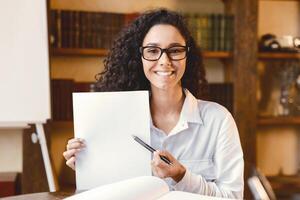 Sign Here. Young smiling confident woman wearing eyeglasses pointing to a blank empty sheet of paper, holding a pen, looking and showing it at camera. Presentation, Agreement, Business Concept. photo