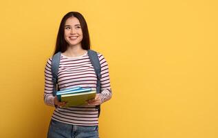 A cheerful student holds a stack of colorful books while wearing a striped shirt and a backpack. The bright yellow background enhances the positive atmosphere of learning and education, copy space photo