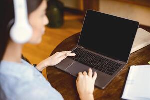 People And Technology Concept. Over the shoulder view of woman in headphones sitting at table and using laptop with black empty screen for mock up, typing on keyboard, studying working online at home photo