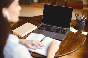 Watching Webinar. Over the shoulder view of lady in earbuds sitting at table with open notebook, using laptop with black empty screen for mockup template, having virtual meeting, call conference photo