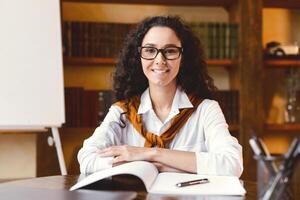 Portrait of positive confident curly woman in eyeglasses sitting at desk with open notebook, bookshelves and board in the blurred background. Lady in white shirt smiling and ready for online tutoring photo
