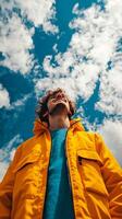A joyful man smiles radiantly, looking up towards a vibrant blue sky filled with fluffy white clouds. He is wearing a bright yello photo