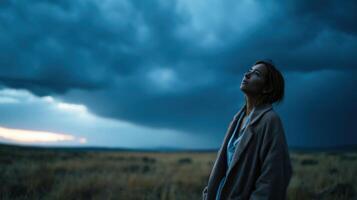 A woman stands in an open field, looking up at a dramatic sunset sky. The vastness of the landscape and the stormy clouds create photo