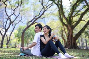 Young asian couple is having a break after happy dating exercise while sitting on the yoga mat in the public park for healthy lifestyle and love usage photo
