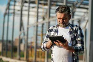 Information in the digital tablet, reading. Senior worker is near the unfinished greenhouse, working photo