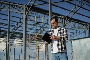 Wireless device, holding digital ,,,tablet in hands. Senior worker is near the unfinished greenhouse, working photo