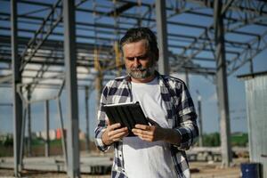 Wireless,, device, holding ,digital tablet in ,hands. Senior worker is near the unfinished greenhouse, working photo