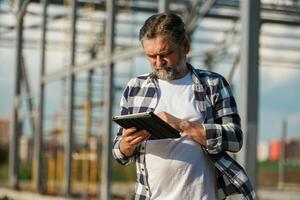 Information ,in the digital tablet, reading... Senior worker is near the unfinished greenhouse, working photo