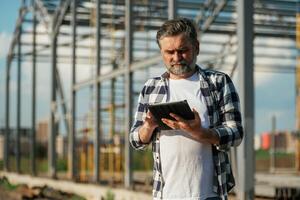 Information in the digital table,,t, reading. Senior worker is near the unfinished greenhouse, working photo