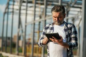 Information in the digital tablet, ,,reading. Senior worker is near the unfinished greenhouse, working photo