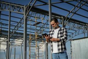 Wireless, device, holding digital tablet in hands. Senior worker is near the unfinished greenhouse, working photo