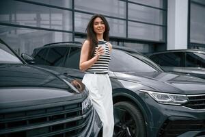 Holding, cup of coffee. Attractive young woman is outdoors near her brand new car photo