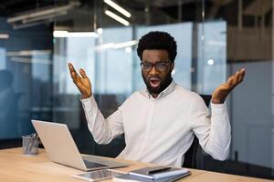Portrait of upset and disappointed man inside office. Businessman looking nervously at camera while working with laptop at workplace. photo