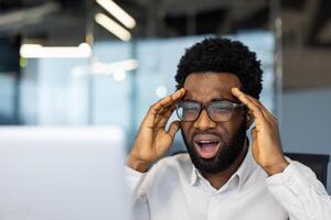 A Black man in glasses reacts to bad news on his computer, with an expression of frustration and hands to his temples photo