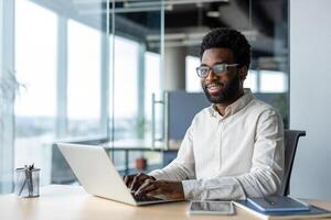 An African American man with glasses working on a laptop in a modern office with a bright window view, showing a happy and productive work environment. photo