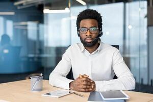 An african-american man wearing a headset sits in an office setting, looking at the camera with a serious expression, ready to assist. photo