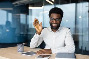 Smiling, well-dressed businessman with glasses waving his hand in an office setting, greeting during a virtual meeting or call. photo