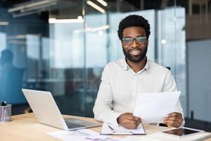 Smiling professional with glasses holds documents while working at his office desk. He looks at the camera with confidence and ease. photo