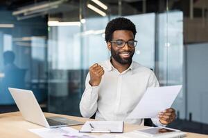 An african american man sits at his desk, ecstatic, holding papers, laptop, and a tablet are on the table. He appears joyful and has an expression of achievement. photo