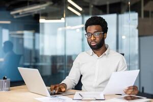 A focused professional balances reviewing documents and working on his laptop in a modern office setting, showcasing productivity and diligence. photo