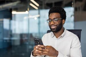 A smiling man wearing glasses looks at his phone while sitting in a modern office. It seems he is reading or watching something interesting. photo