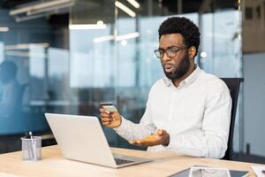 A perplexed African American man in glasses encounters issues while attempting an online transaction using his credit card and laptop in a modern office. photo