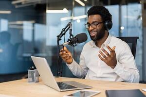 A smiling African-American man with glasses records a podcast in a modern office setting. He is using a microphone, headphones, and a laptop. photo