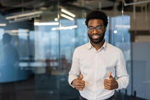 Smiling african american man in eyeglasses gives a thumbs up in an office environment, representing success and approval. photo