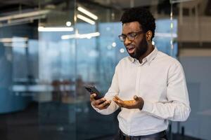 An African American man with glasses looks shocked at something he sees on his phone while in an office setting. photo