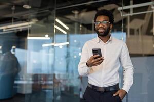 Successful businessman with phone in hands standing inside office at workplace. Man smiling contentedly looking out window, financier investor, using app on smartphone. photo
