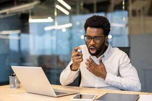An african american man in an office uses an inhaler, potentially experiencing an asthma attack while working on a laptop. photo