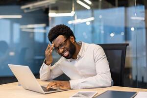 An African-American man working at his desk is shown with a stressed expression. He is experiencing a headache and appears overwhelmed while using his laptop. photo