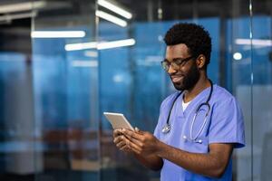 Young successful experienced doctor standing inside the clinic with a tablet computer in his hands. The doctor reviews patients' data and communicates with clients remotely. photo
