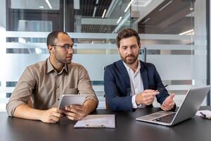 Two business colleagues collaborate, using a laptop and tablet to review documents and discuss ideas at their office table. photo