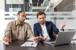 Two businessmen sitting at a desk, looking concerned while discussing something in front of a laptop and tablet in an office setting. photo